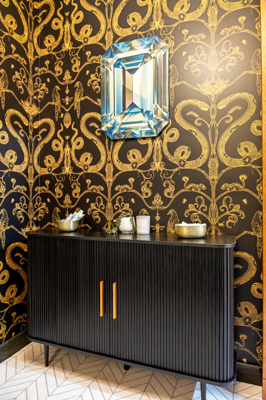 Elegant black cabinet against gold and black snake-patterned wallpaper with a blue gem artwork.