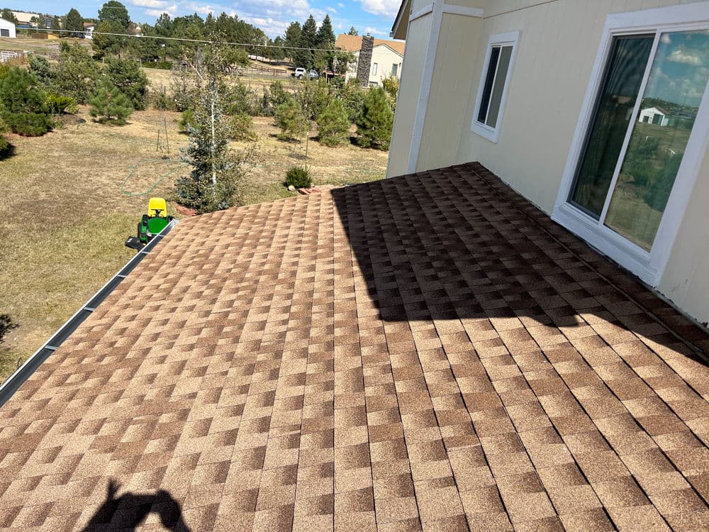 Brown shingle roof viewed from above with green trees and residential area in the background.