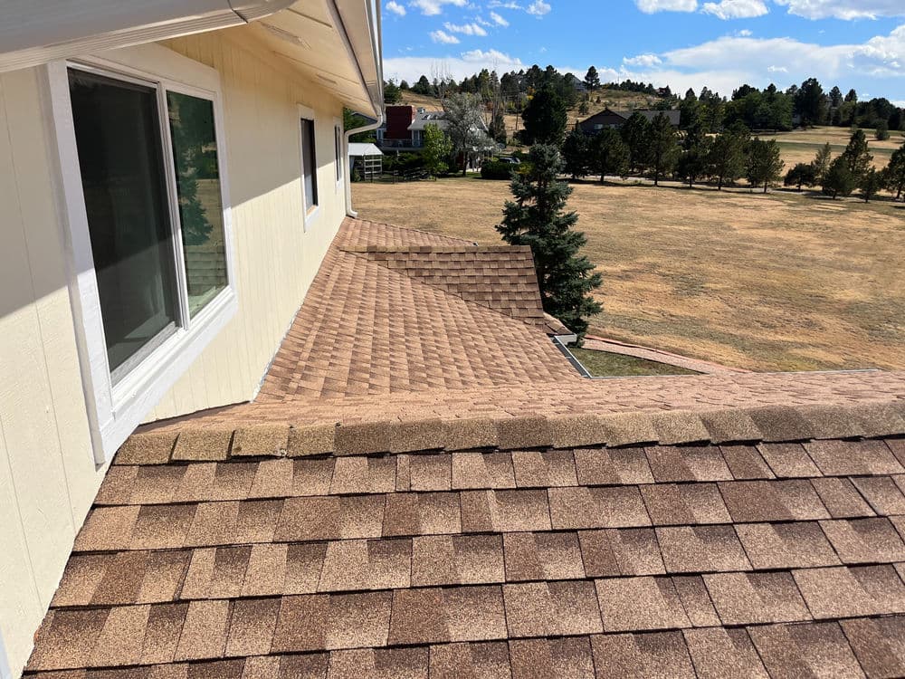 Residential roof with asphalt shingles, overlooking grassy landscape and pine trees.